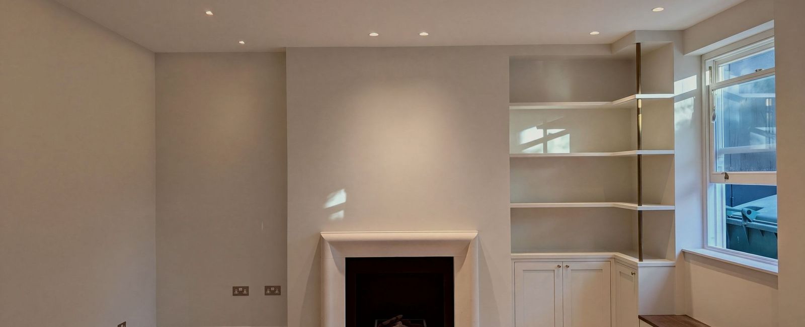 A living room with a stone fireplace surround, built-in alcove shelving and cupboards to the right with a brass upright, a window seat and a herringbone floor