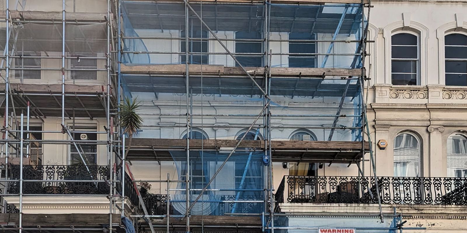 A stucco fronted London terrace under full scaffold and blue sheeting during major works, with the unscaffolded neighbouring houses to the right