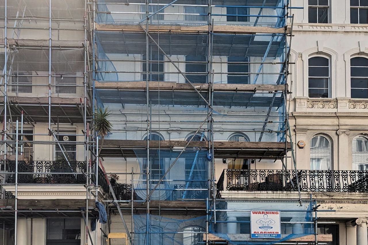 A stucco fronted London terrace under full scaffold and blue sheeting during major works, with the unscaffolded neighbouring houses to the right
