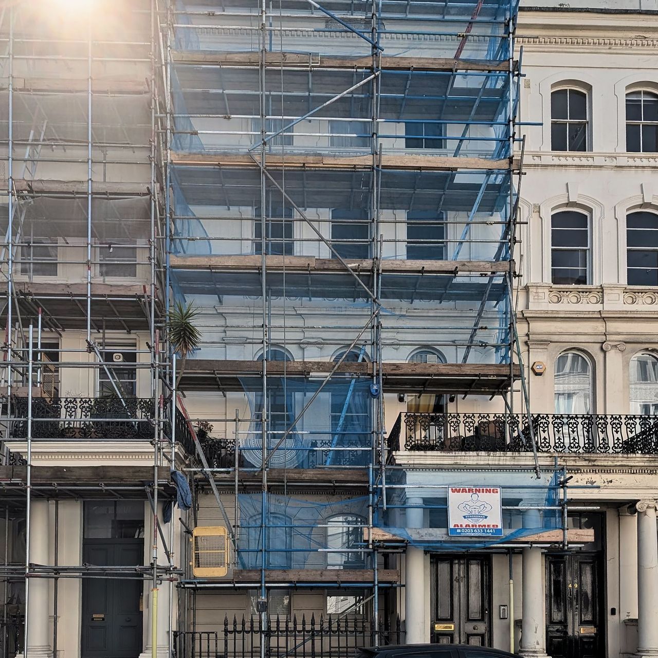 The four storey stucco terrace under full scaffold with blue debris netting, seen from the pavement opposite