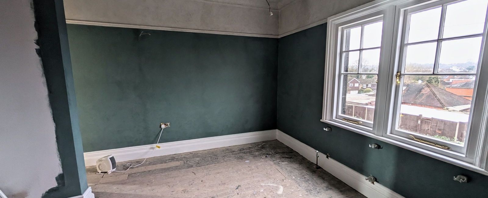 A deep teal room with a chimney breast, white cornice and two sash windows, with the floor still bare and protection to the boards