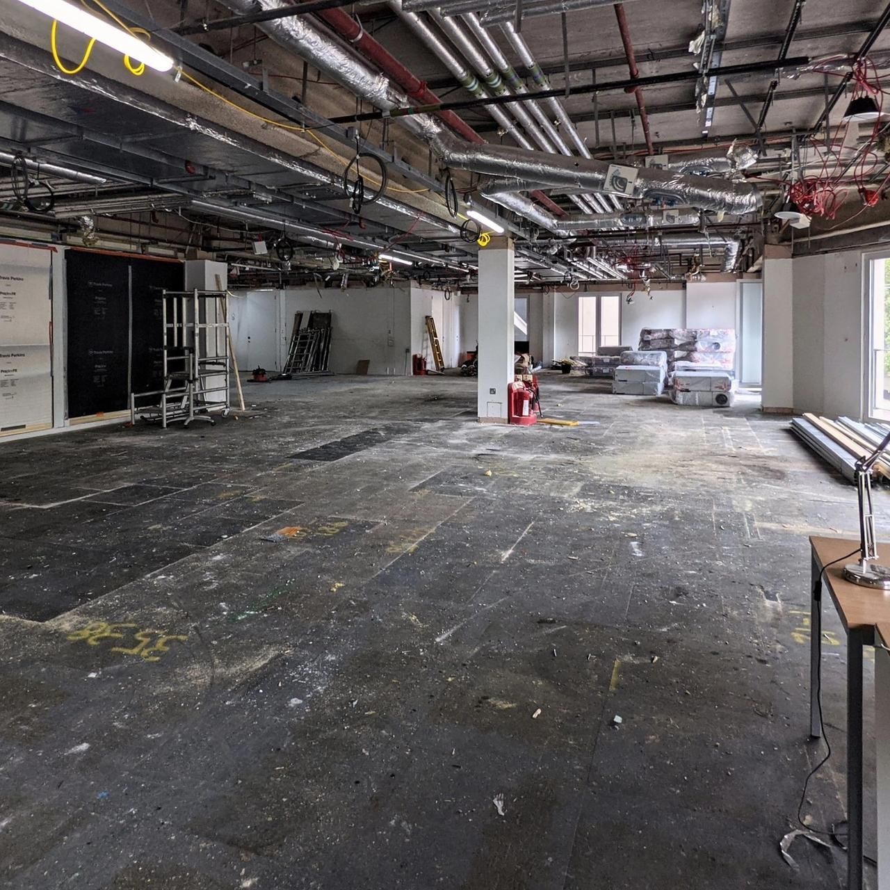 Stripped office floor with plans on a trestle, a desk lamp and windows to the park