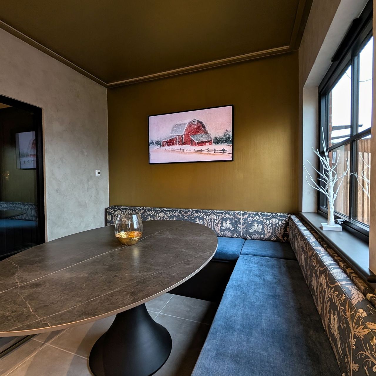 A dressed dining area with an oval dark table, patterned upholstered banquette seating, an olive gold wall and a window to a garden