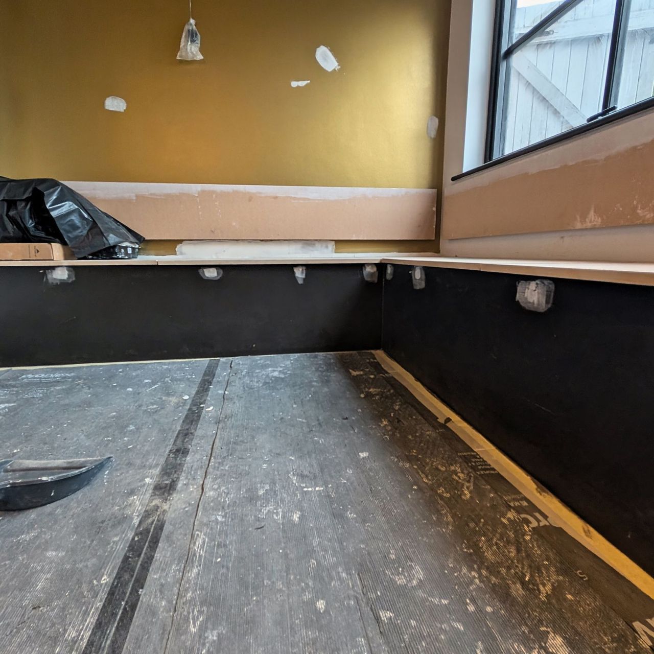 An olive gold upper wall over a dark lower wall, a bench substrate along the window and a scraper left on the protected floor