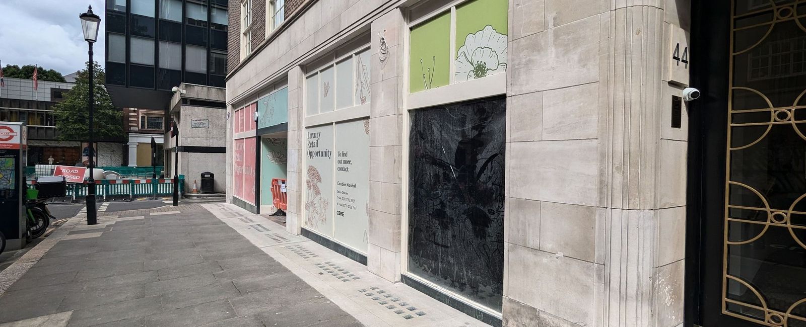 A vacant retail unit on a pale stone elevation before work, the shop windows blanked with letting vinyls and one bay painted out dark