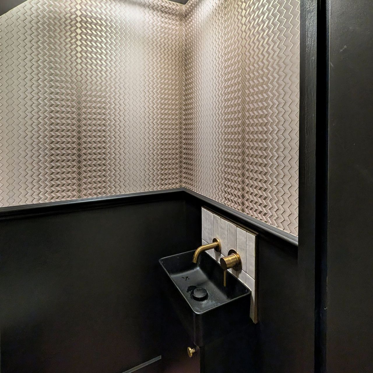 An embossed chevron wall covering above dark painted panelling, with a black wall hung basin and brass tap and a dark panelled door
