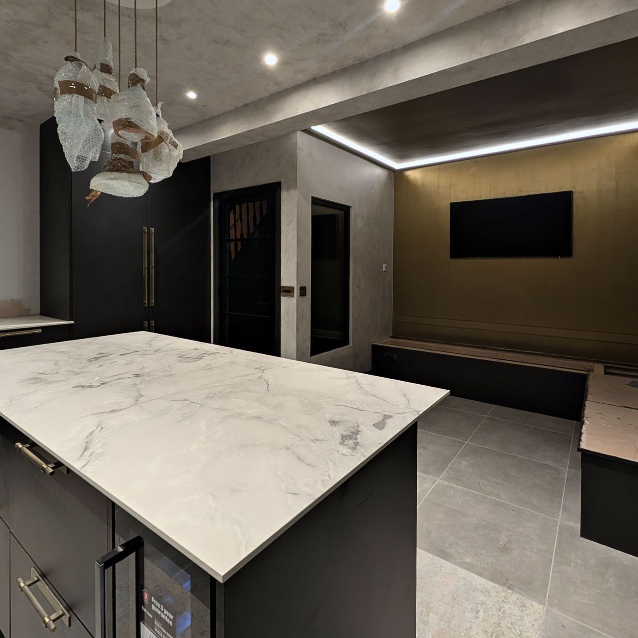 The marble island from the far end with glass pendants above, dark units to one side and the gold wall and banquette beyond
