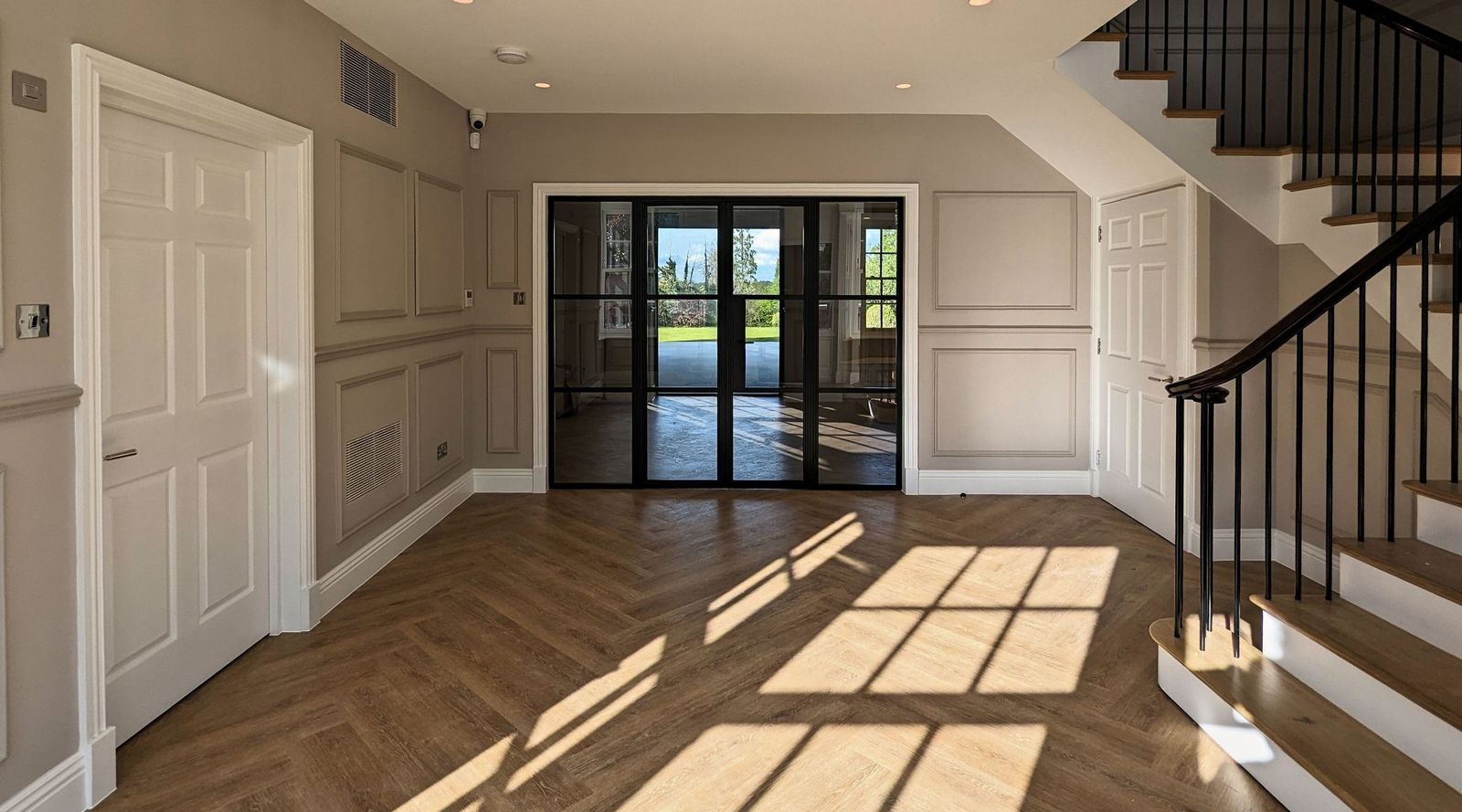 Panelled hall of a family house in Cuffley looking through black glazed steel doors to the garden, with afternoon light across a herringbone floor