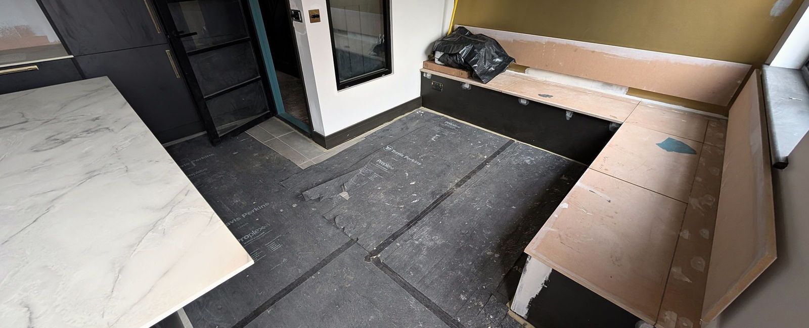 A white marble slab resting on trestles beside a plywood banquette carcass, dark units to the left and protection over the floor