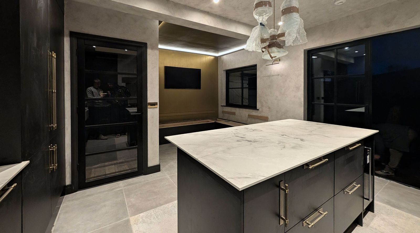A dark fitted kitchen with a white marble island, glass pendant lights over it, softly mottled pale plaster-effect walls and an olive gold feature wall at the far end.