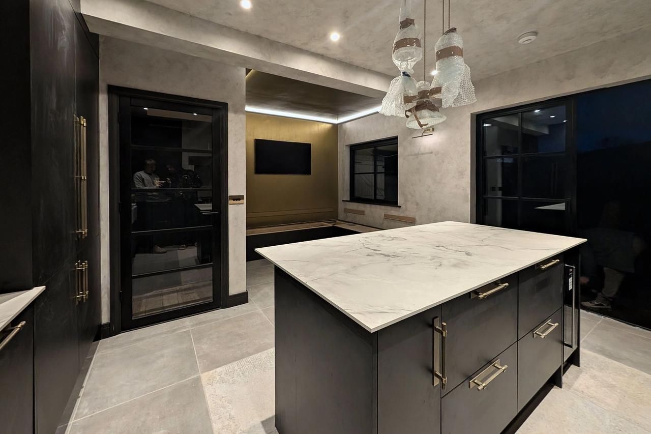 A dark fitted kitchen with a white marble island, glass pendant lights over it, softly mottled pale plaster-effect walls and an olive gold feature wall at the far end.