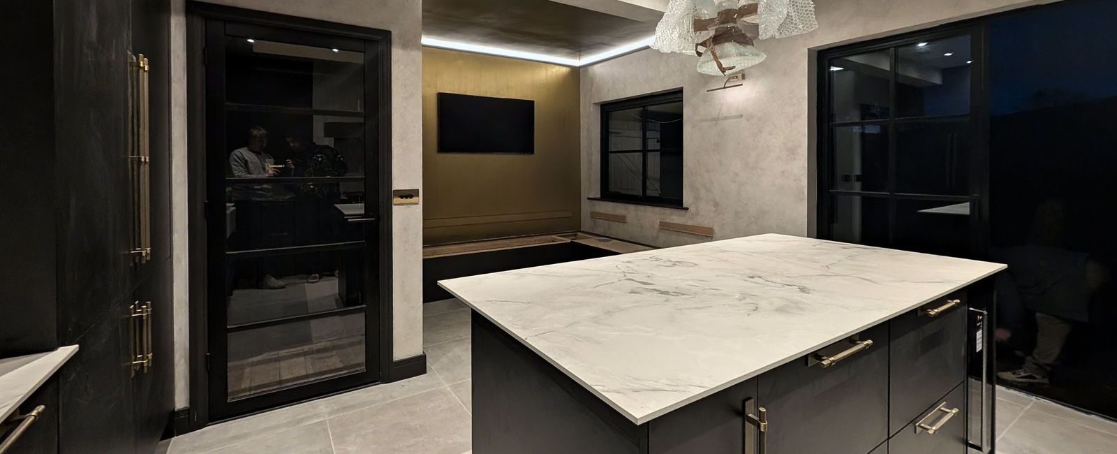 A dark fitted kitchen with a white marble island, glass pendant lights, softly mottled pale walls and an olive gold wall at the far end