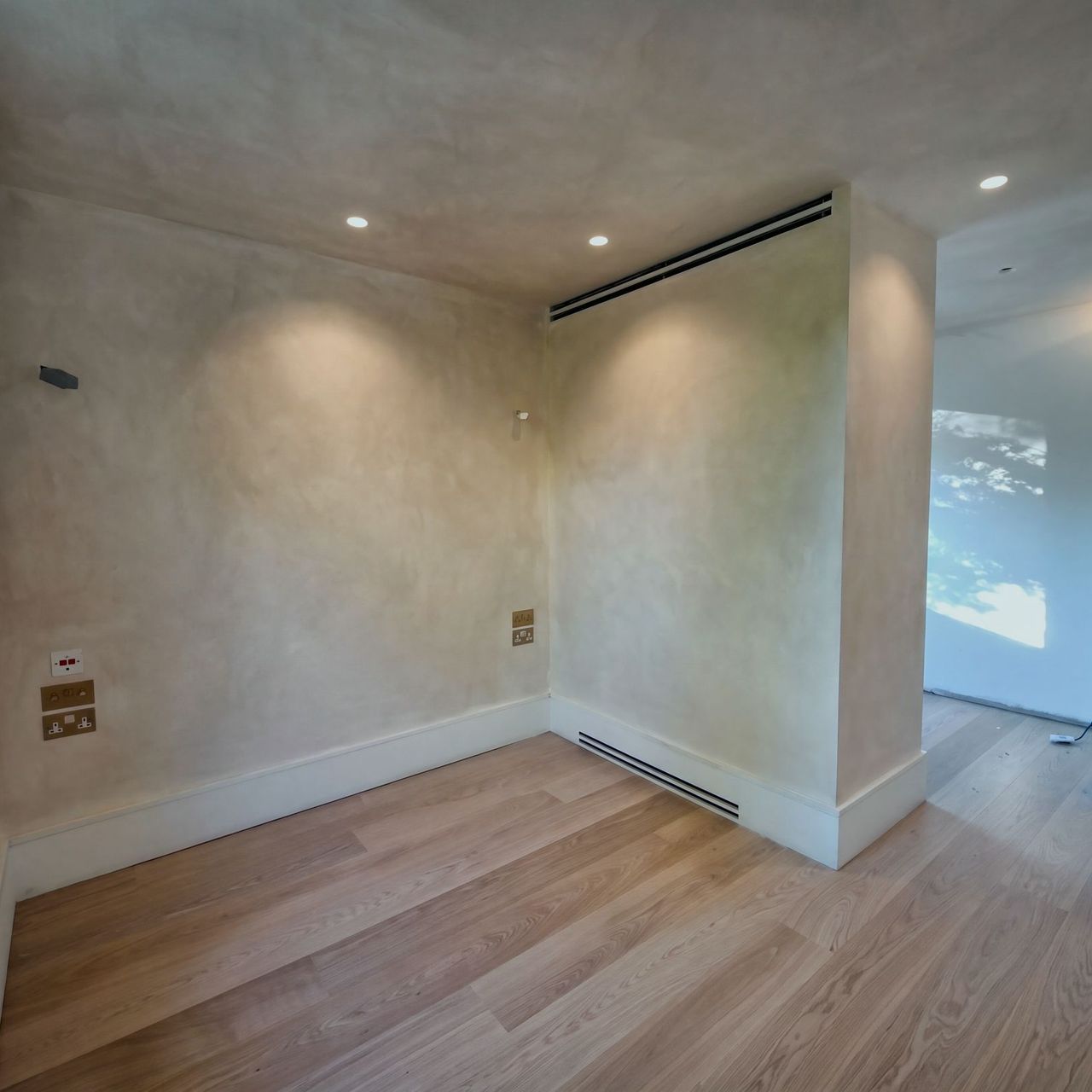 A limewashed corner with a window sill to the left, a vent set into the painted skirting and a pale plank floor running through to the next room
