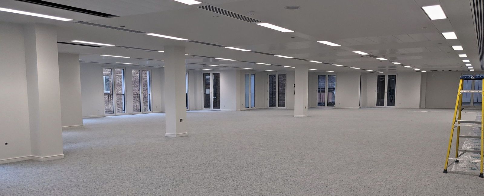 Finished open plan office floor with white columns, a grey carpet and windows along both long walls