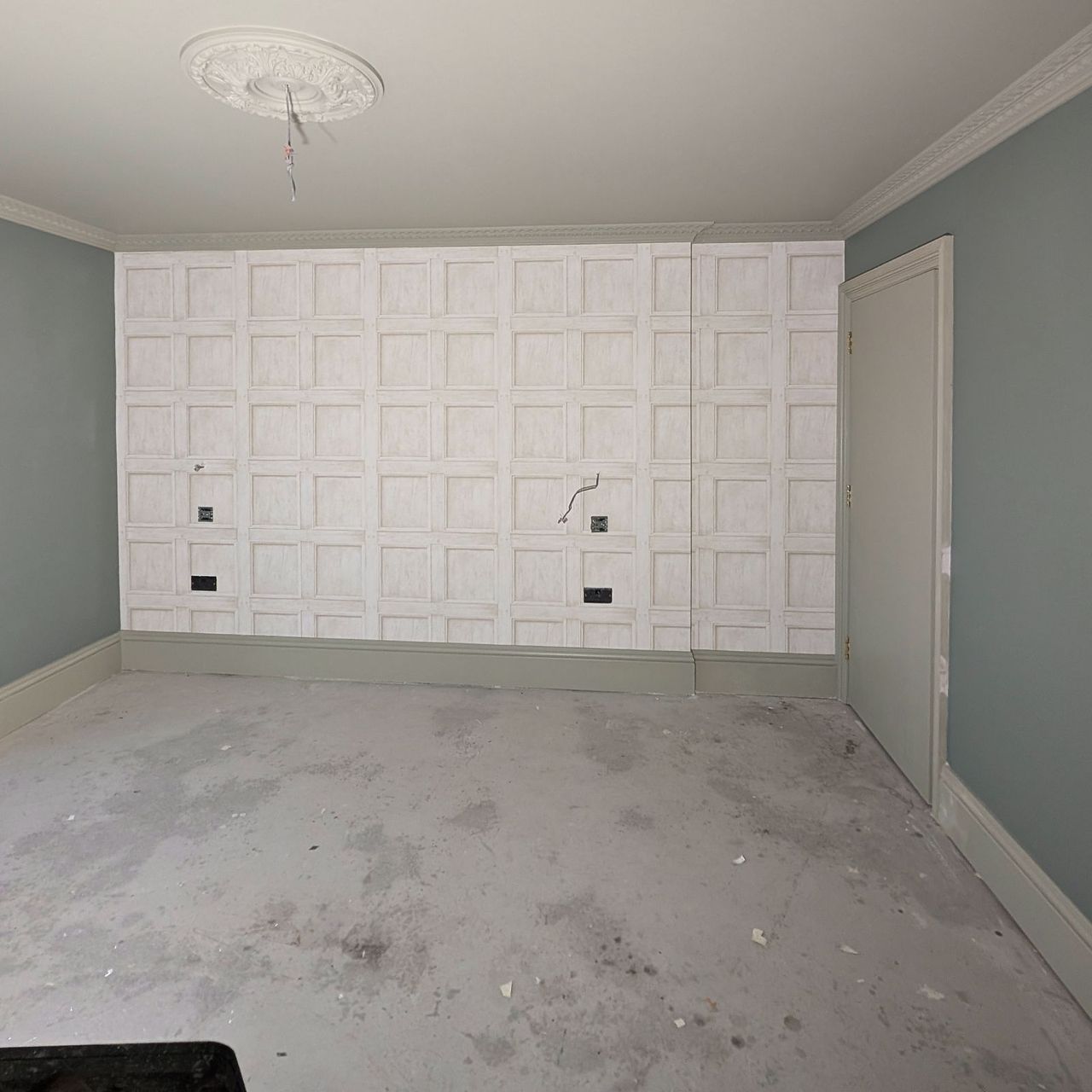 A feature wall papered in a square panel pattern across the end of a room with pale sage walls, a ceiling rose and a bare floor