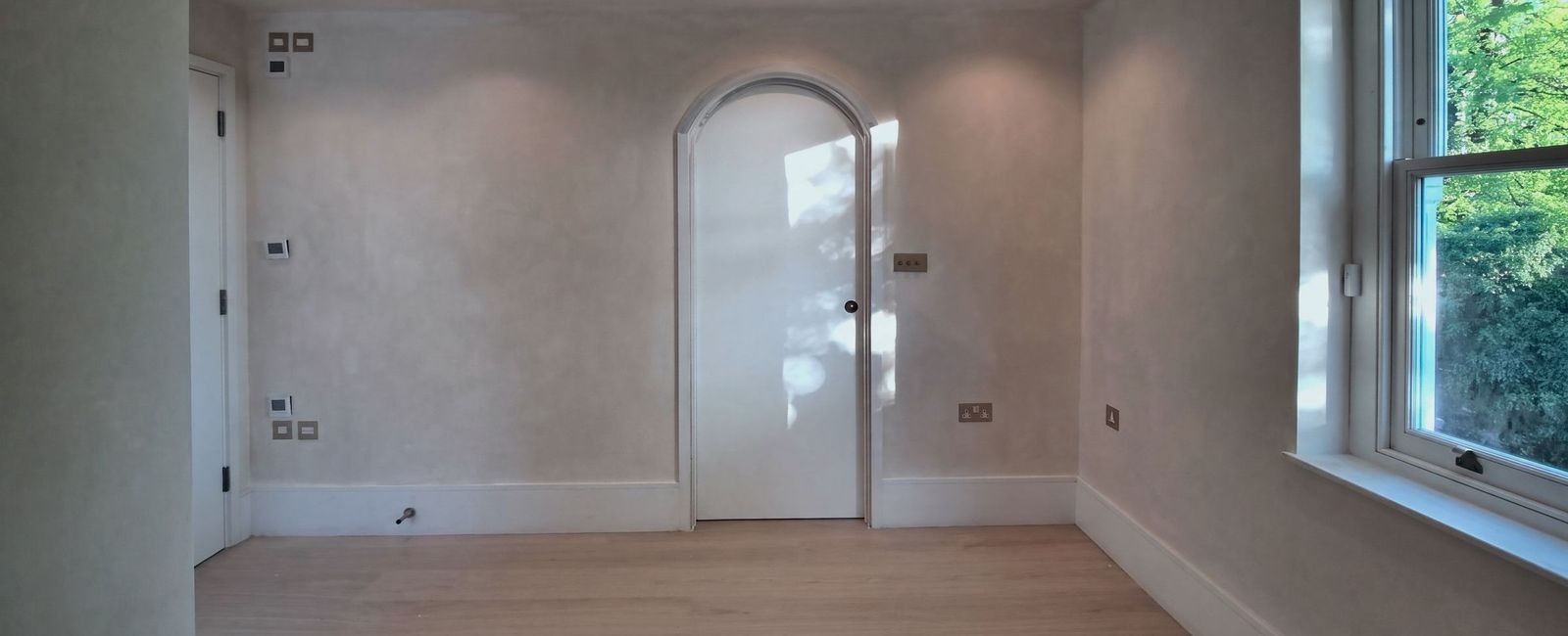 A limewashed room with a white arched door at the centre catching the sun, brass sockets, a pale plank floor and a sash window to the right