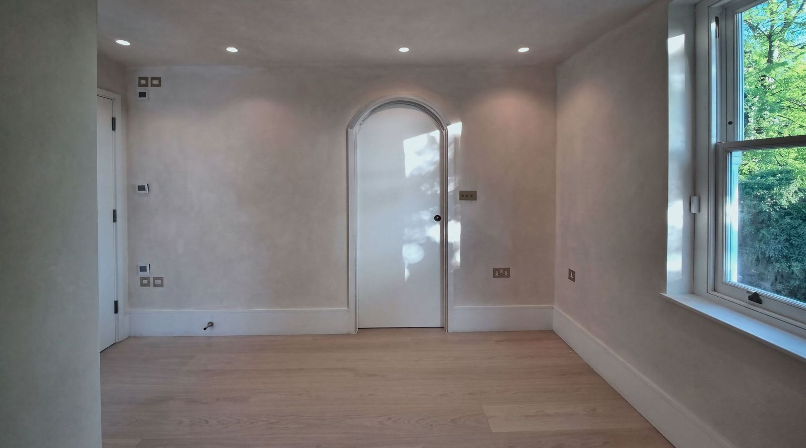 A limewashed room with a white arched door at the centre catching the sun, brass sockets, a pale plank floor and a sash window to trees on the right.