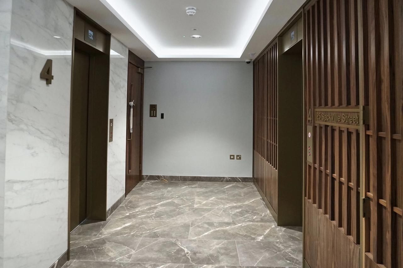 Lift lobby of a Mayfair office building with marble walls, a brass lift door, walnut slatted screens and a lit ceiling cove