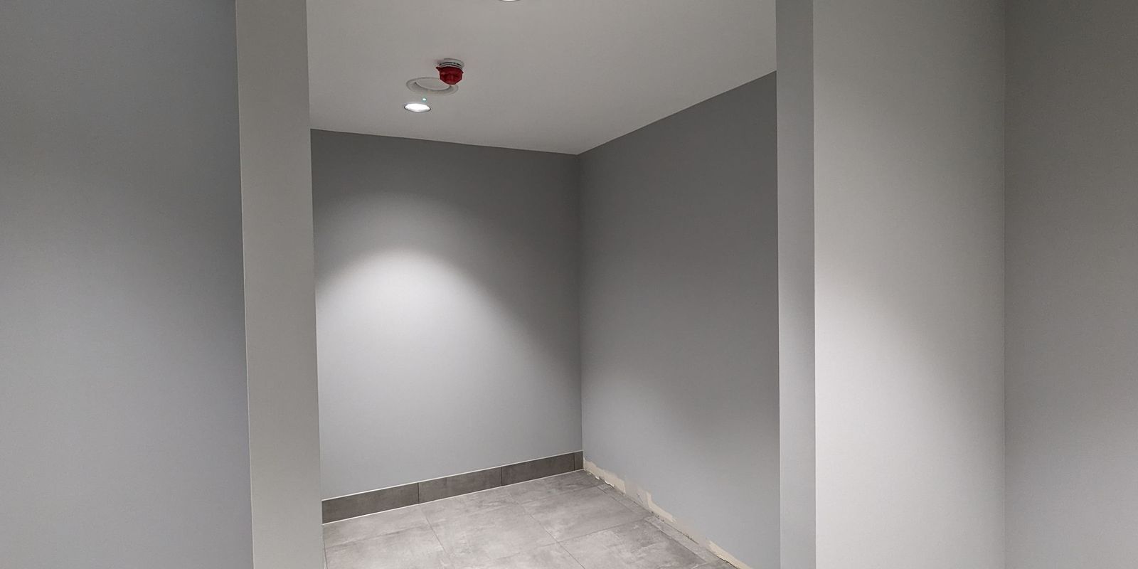 A finished communal area in grey, with a recessed downlight, a red ceiling sounder and a large format tiled floor
