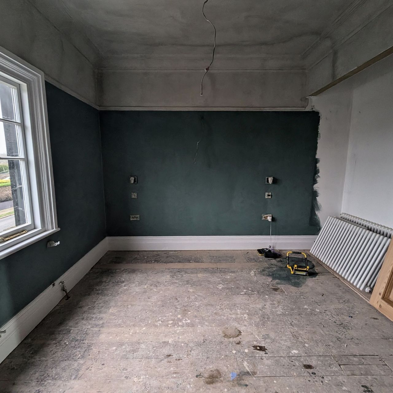 A room in deep teal with white cornice and skirting, sash windows to the left, a bare timber panelled door leaning against the wall