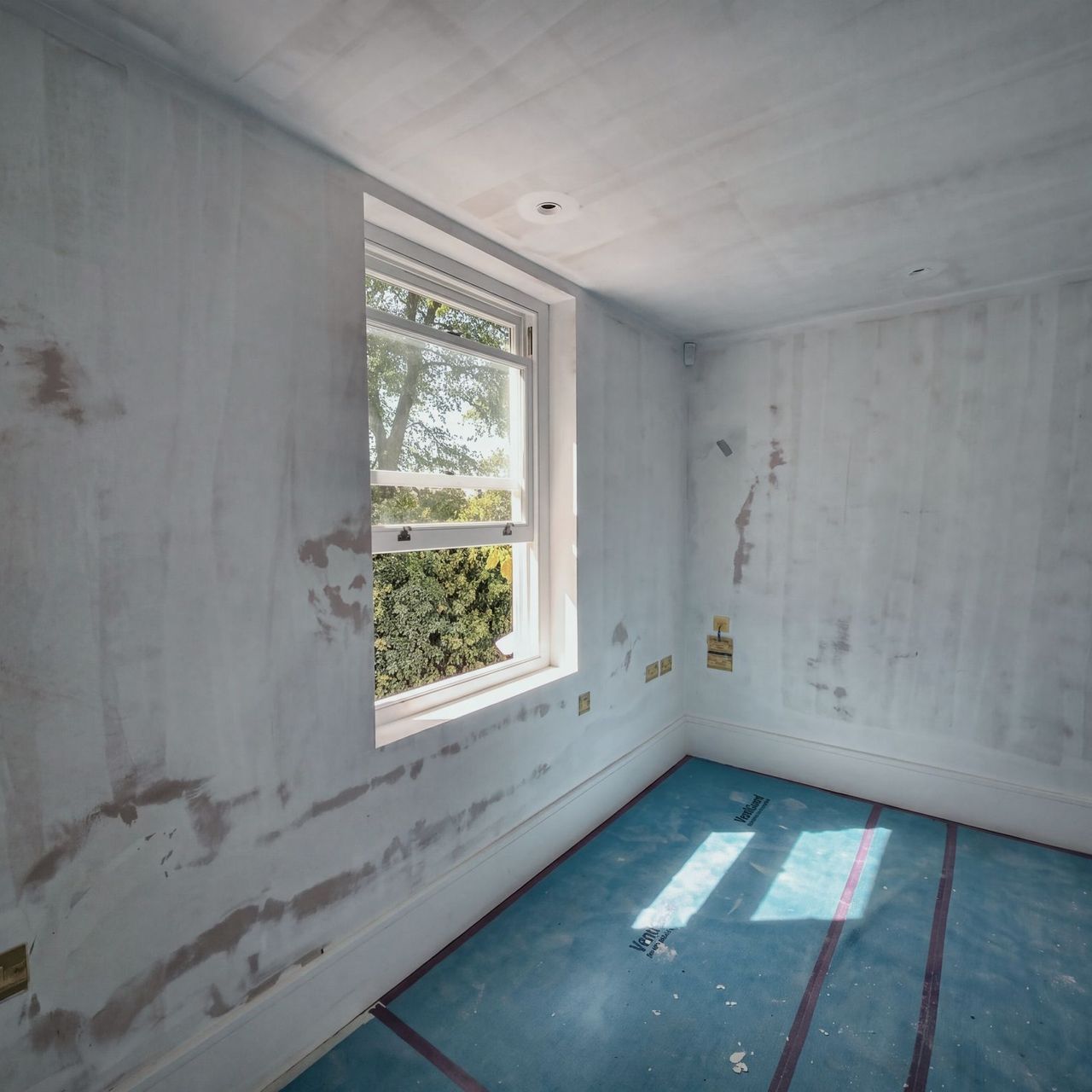 A room in patched plaster with a sash window looking into trees, brass sockets and blue protection with sunlight falling across it