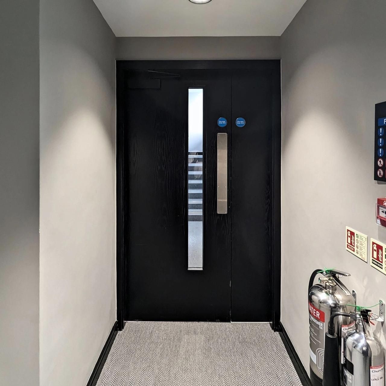 Black flat entrance door with a vertical glazed panel in a grey painted lobby with fire safety signage