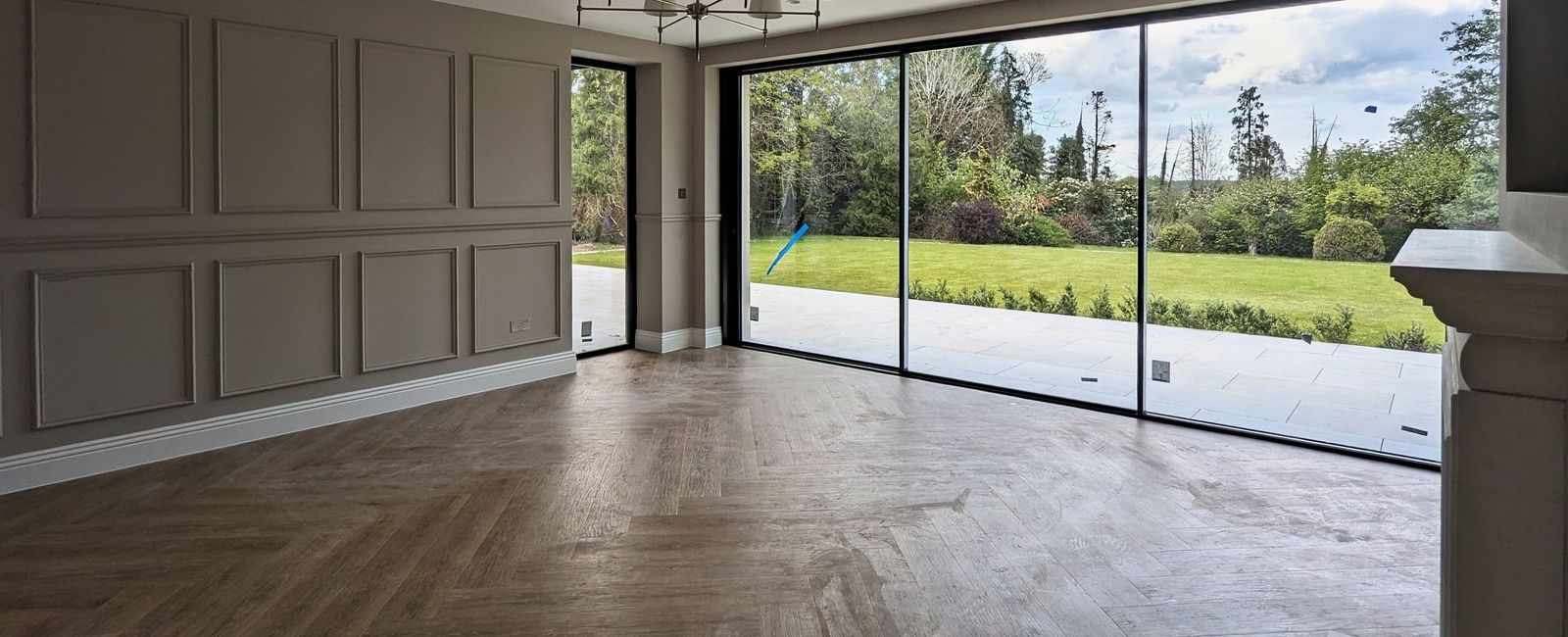Living room panelling running to full-width sliding glass doors onto a lawn, herringbone floor in the foreground