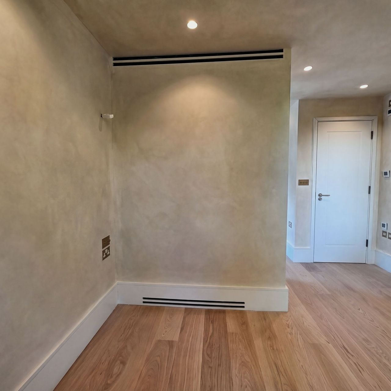 A limewashed room with a vent in the skirting, a white panelled door to the right opening to a landing and a pale plank floor under downlights