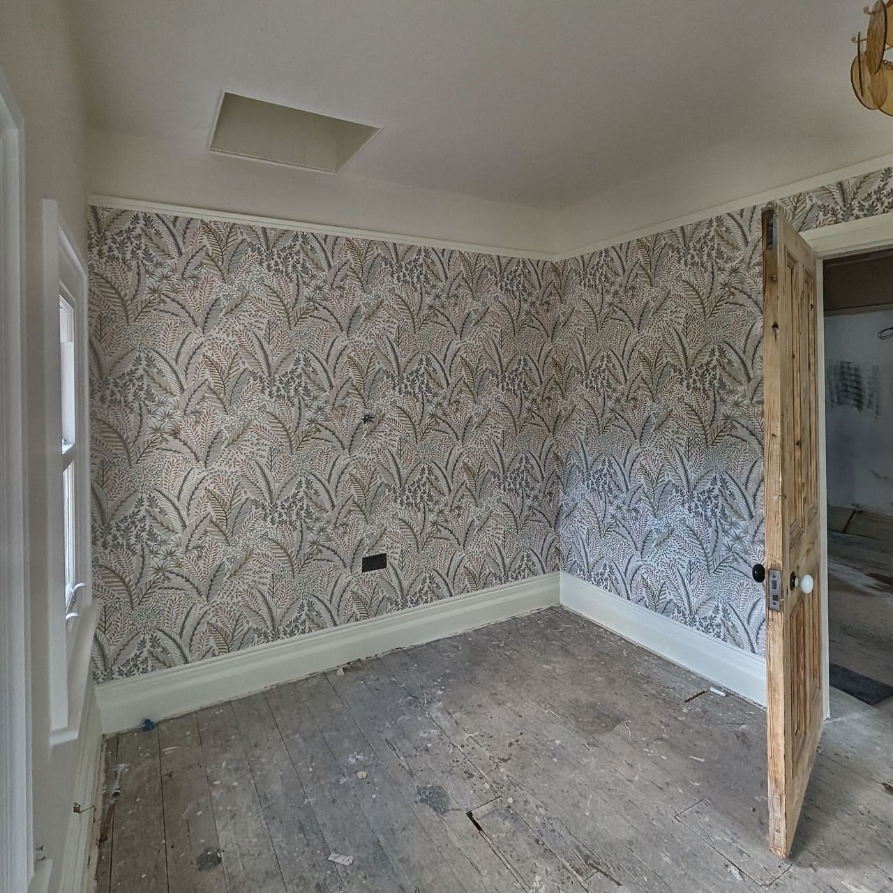 A wall papered in a pale grey pattern beside an open stripped timber door, with a sash window to the left and a bare floor