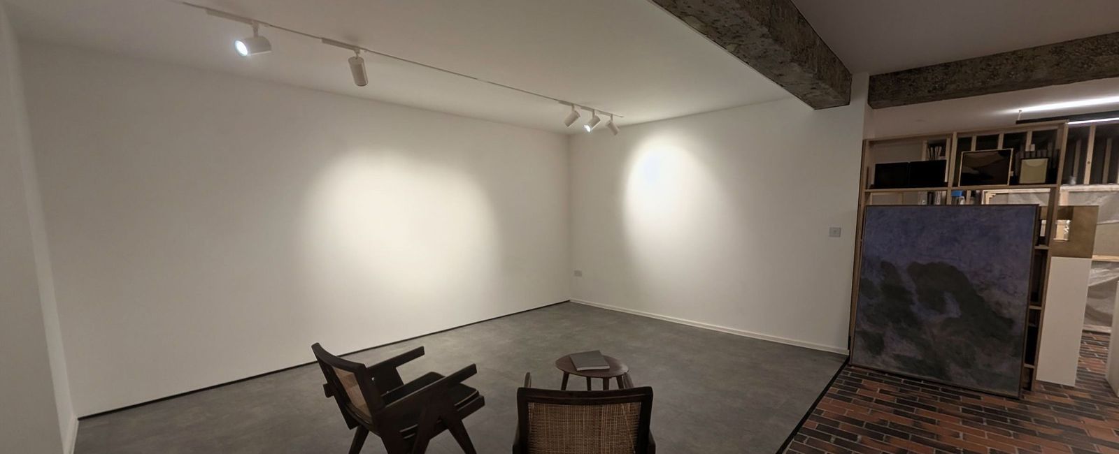 The lower gallery finished in matt white with track lighting, an exposed structural downstand left unfinished and a brick paviour and screed floor