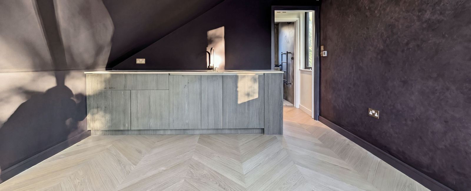 An aubergine sloped ceiling over a pale timber fronted counter with a tap, a doorway through to a lit landing and a chevron floor