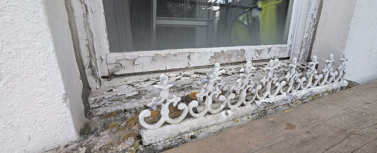 A sash window sill with a decorative cast iron guard, paint flaking from the sill and the moulding beneath