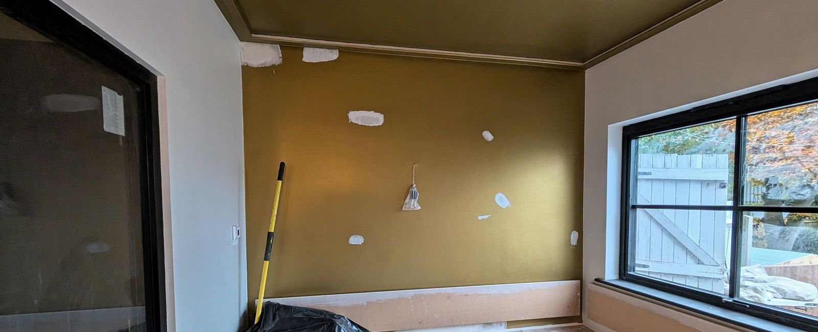 An olive gold wall marked with white filler patches above a dark bench base, black framed glazing to the right and floor protection down