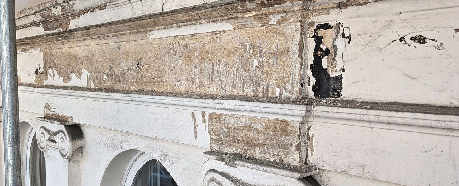 A run of stucco cornice and a console bracket with heavy paint loss and patched repairs, a large black patch of failed render, and two Ionic capitals over arched window heads below