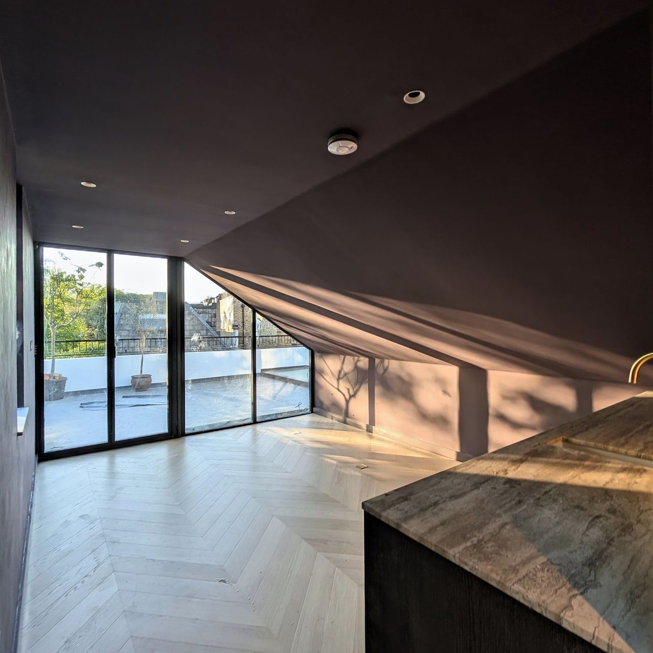 A room with deep aubergine hand-finished walls and a sloped ceiling, full height glazing out to a roof terrace, a stone topped counter to the right and a chevron floor