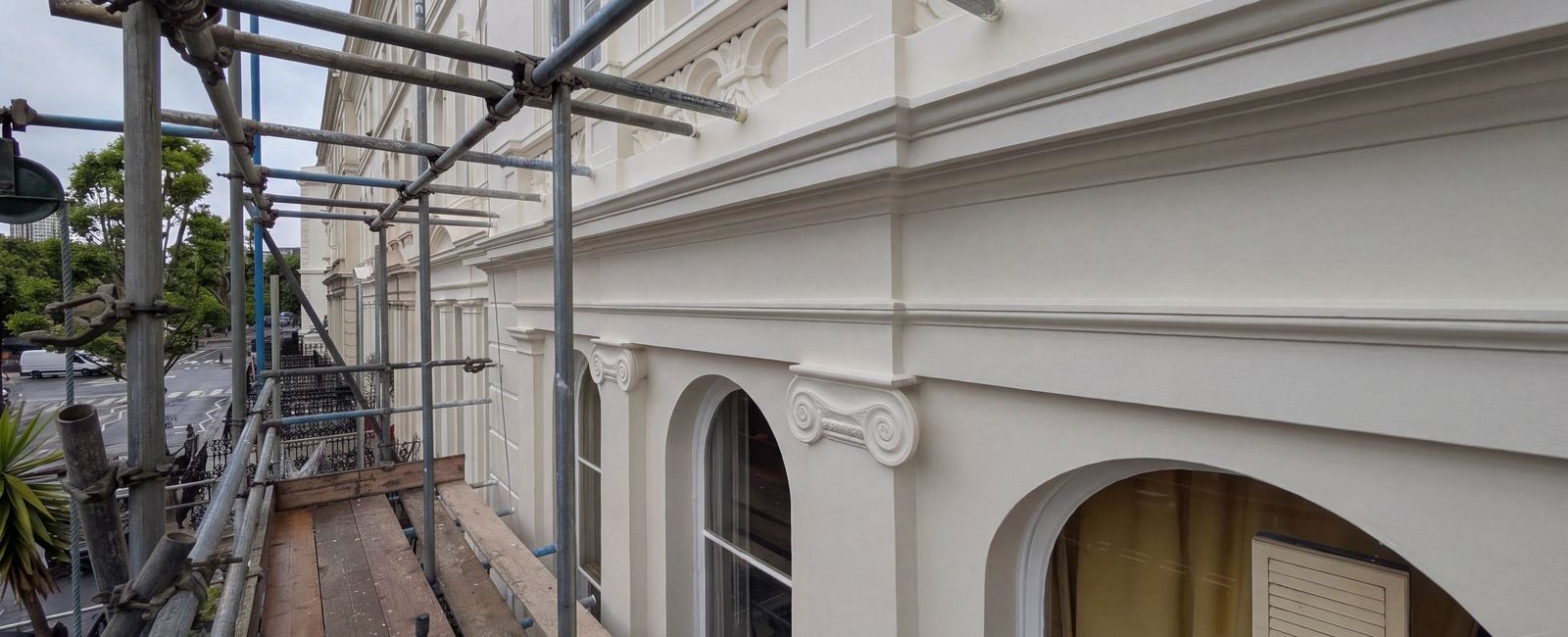 The finished cream stucco elevation seen along the scaffold lift, with the cornice, the enriched band, the Ionic capitals and the arched window heads all complete
