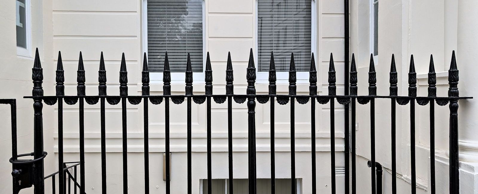 A full width run of black cast iron railings with spear heads and acanthus finials, in front of the finished cream stucco elevation, with sash windows above and the basement area below