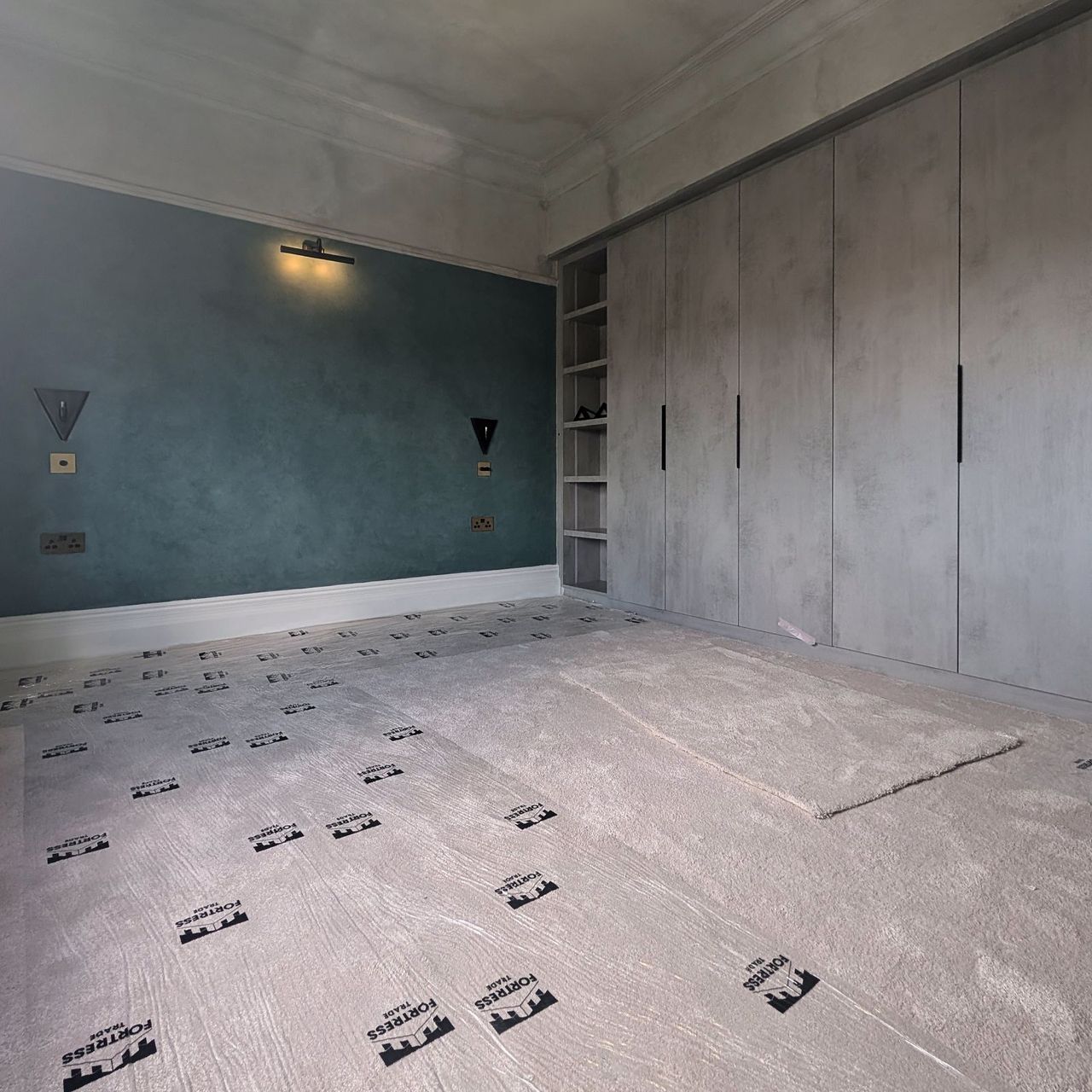 A bedroom with a deep teal wall, wall lights, a radiator and fitted wardrobes in pale grey green to the right, with floor protection down