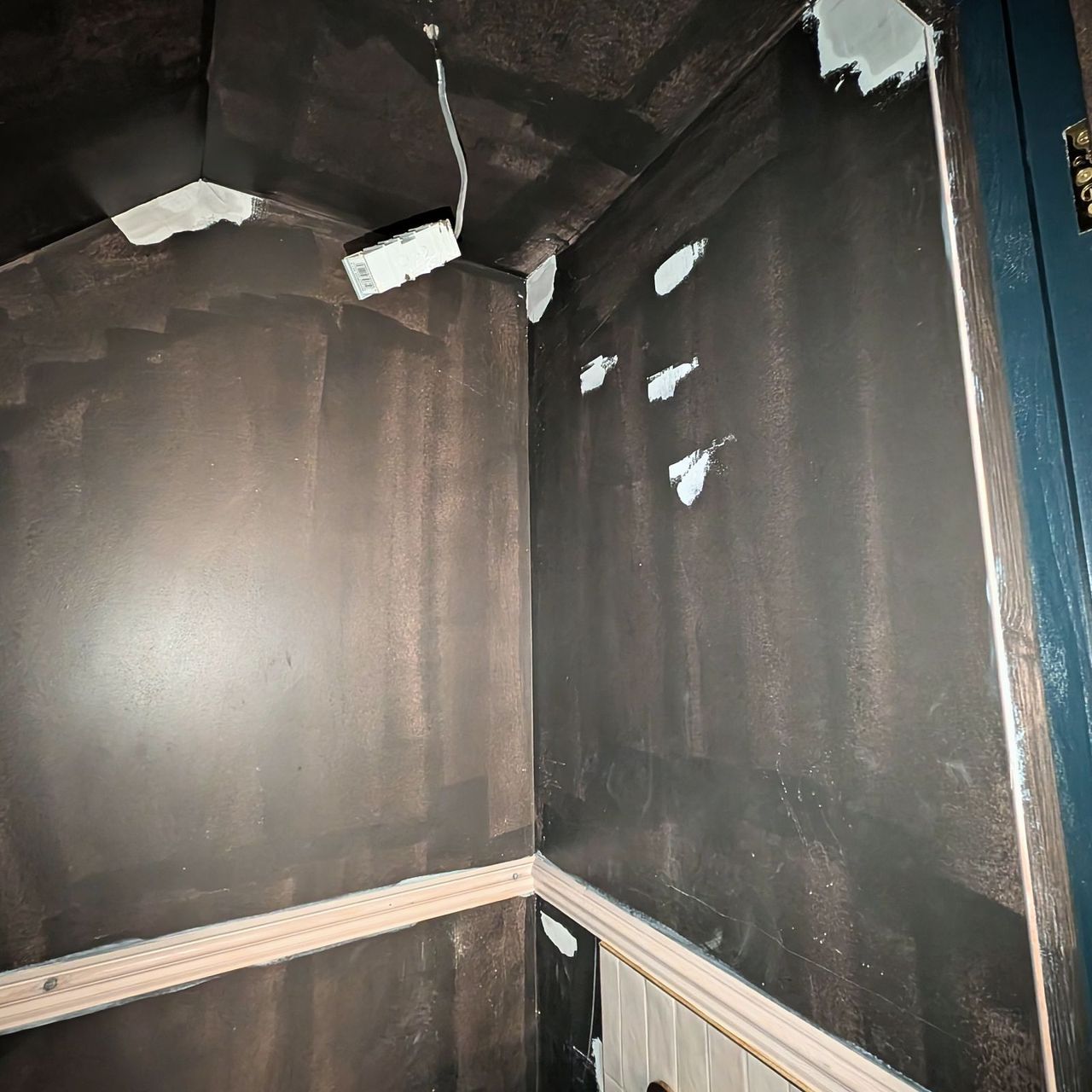 A small room with dark charcoal walls and ceiling dotted with white filler patches, a teal painted door frame and a timber rail across the wall