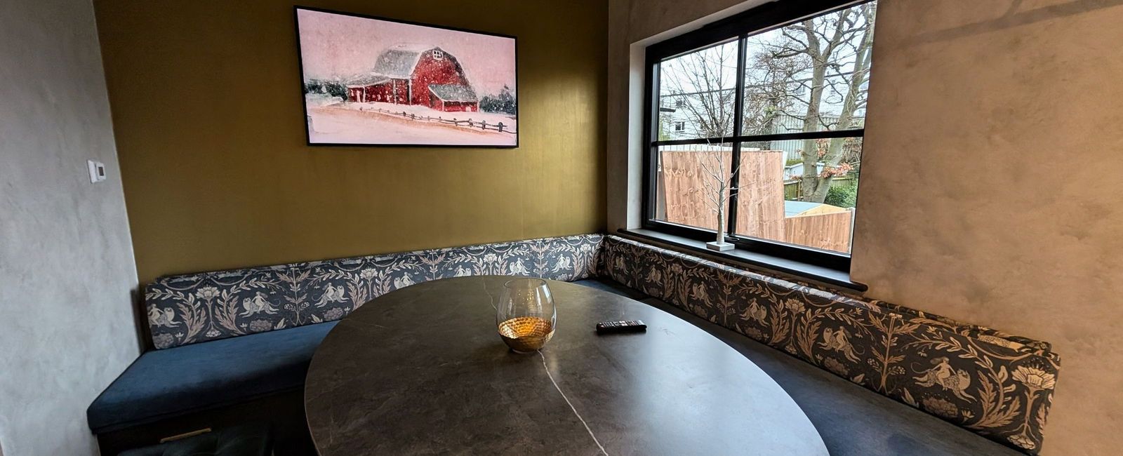 An upholstered banquette running two walls around an oval table, beneath a gold wall with a wall mounted television and a window beyond
