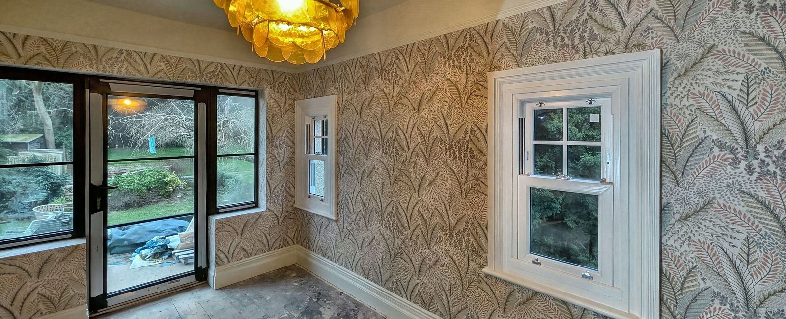 A room papered in a large gold and green leaf pattern with an amber glass tiered pendant, a black framed window and a white sash window