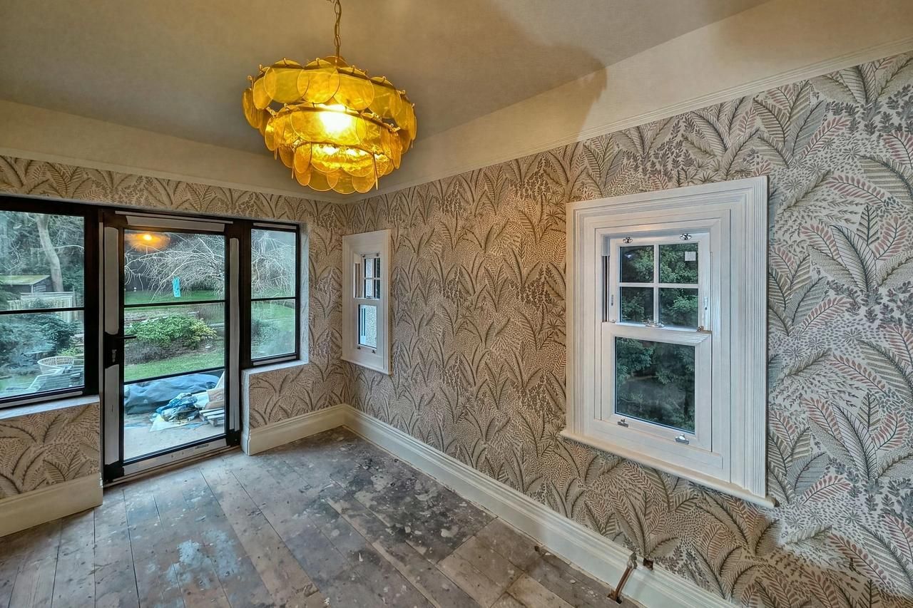 A room papered in a large gold and green leaf pattern, with an amber glass tiered pendant light, a black framed window and a white painted sash window.