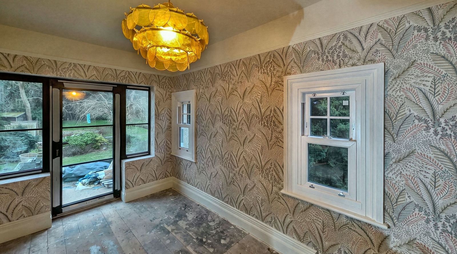 A room papered in a large gold and green leaf pattern, with an amber glass tiered pendant light, a black framed window and a white painted sash window.