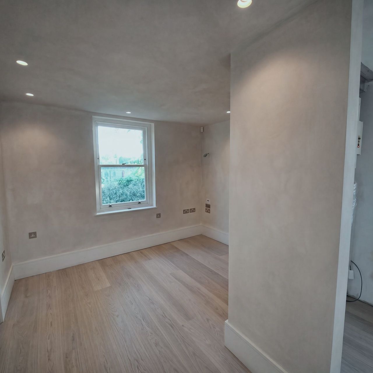 A finished room in pale limewash with an arched opening to the left, a sash window beyond and a pale plank floor under downlights