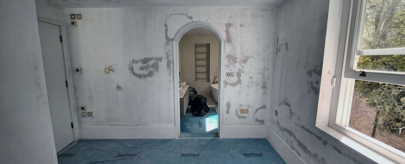 A room in patched bare plaster with a white moulded arched opening through the far wall to a bathroom beyond, and blue floor protection covering the floor