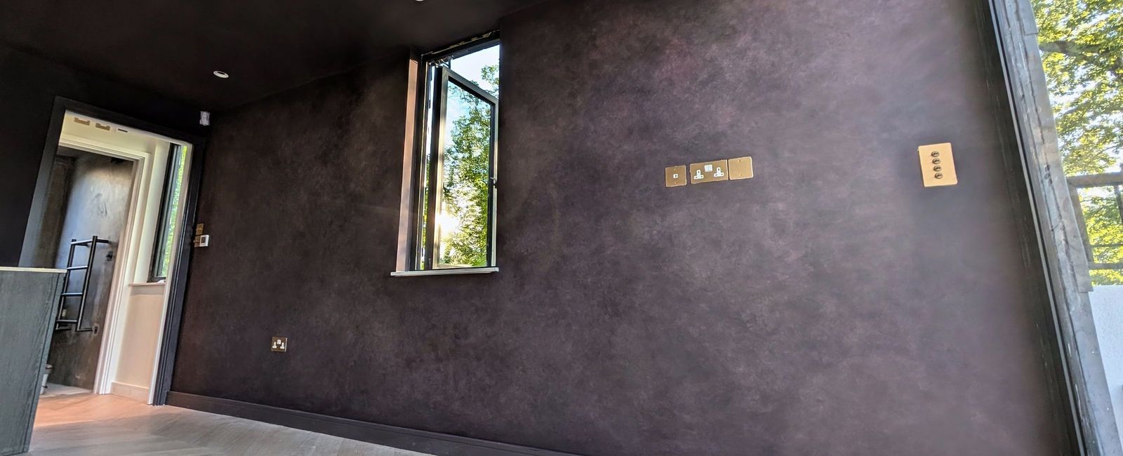 A deep aubergine wall with a mottled hand-applied finish, a casement window to the terrace, brass sockets and switches over a chevron floor