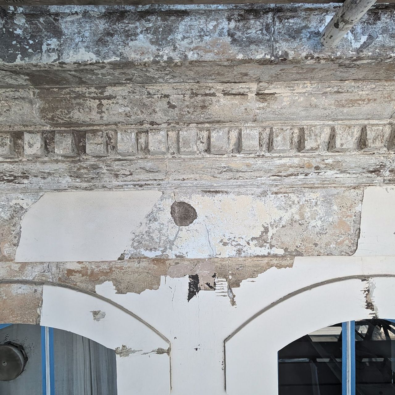 A dentilled stucco cornice with large areas of paint and render missing and bare substrate exposed between the dentils