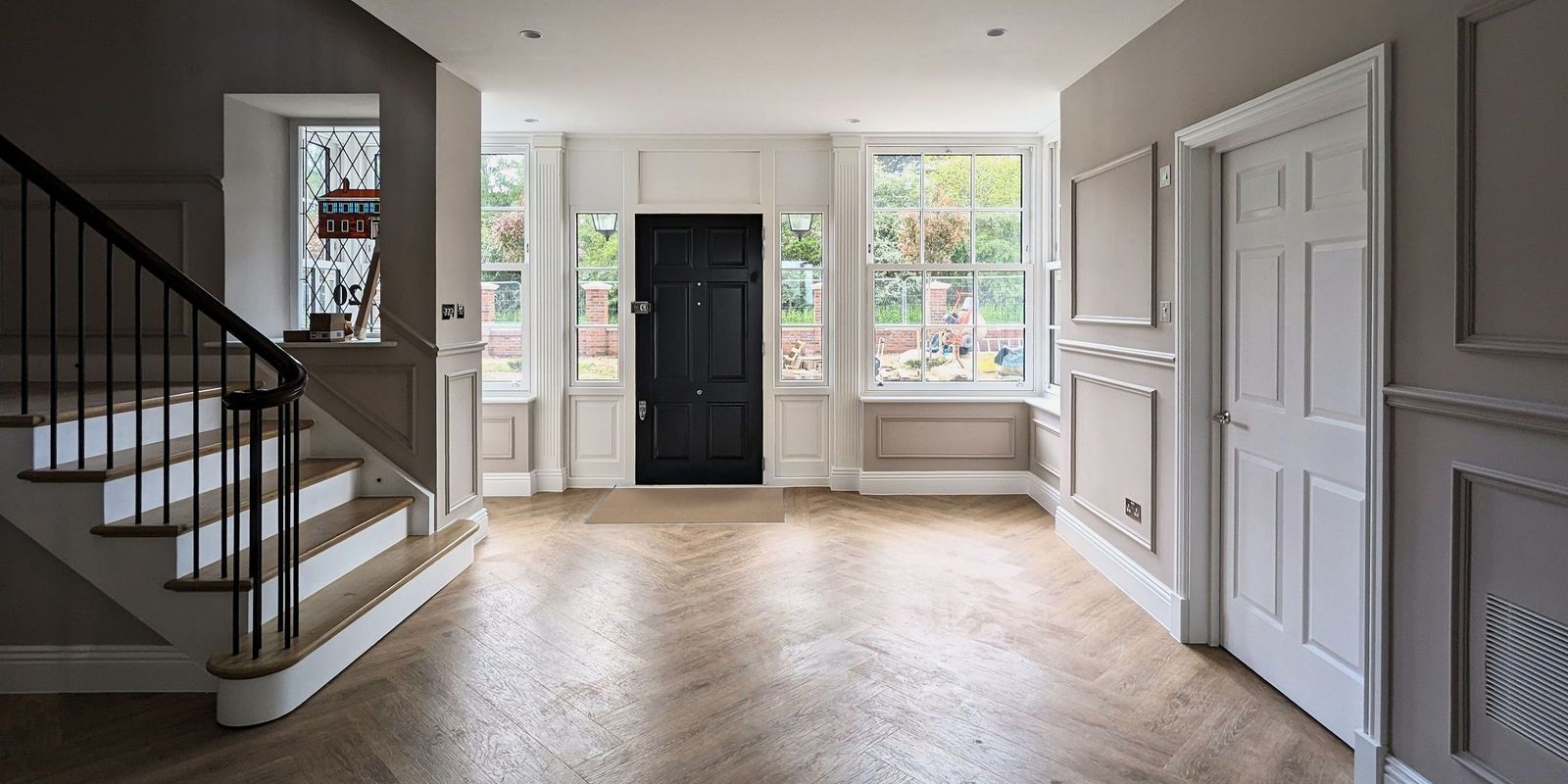 Entrance hall of a period family house with painted wall panelling either side of a black front door, a stair rising to the left and a herringbone floor