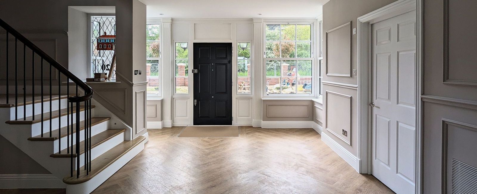 Entrance hall of a family house in Cuffley with painted panelling either side of a black front door, the stair rising to the left and a herringbone floor
