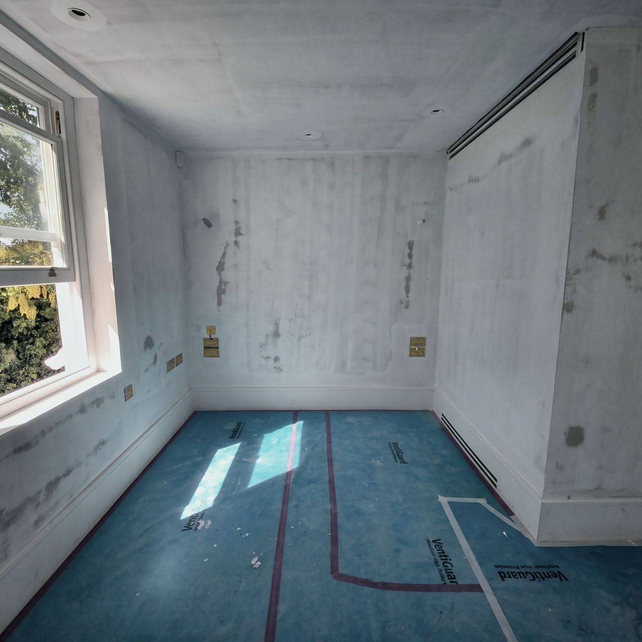 Patched plaster walls around a sash window to the left, brass socket plates, a slim vent above and blue floor protection throughout